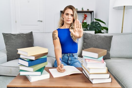 Young beautiful woman writing on notebook around university books with open hand doing stop sign with serious and confident expression, defense gesture