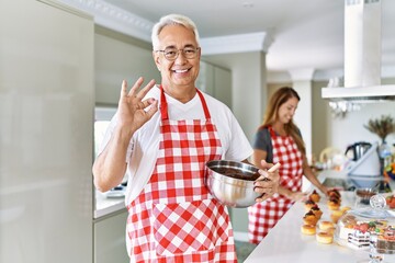 Middle age hispanic couple wearing apron cooking homemade pastry doing ok sign with fingers, smiling friendly gesturing excellent symbol