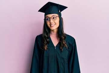 Young hispanic woman wearing graduation cap and ceremony robe looking away to side with smile on face, natural expression. laughing confident.