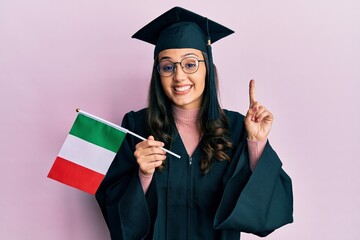 Young hispanic woman wearing graduation uniform holding italy flag smiling with an idea or question pointing finger with happy face, number one