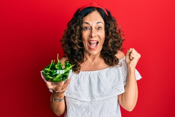 Middle age hispanic woman holding bowl with green peppers screaming proud, celebrating victory and success very excited with raised arm