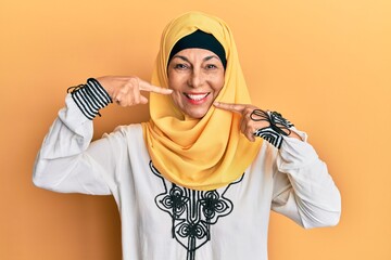 Middle age hispanic woman wearing traditional islamic hijab scarf smiling cheerful showing and pointing with fingers teeth and mouth. dental health concept.