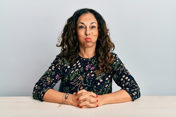 Beautiful middle age woman wearing casual clothes sitting on the table puffing cheeks with funny...