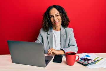 Beautiful middle age woman working at the office drinking a cup of coffee winking looking at the...