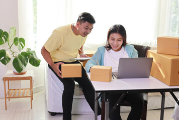 Asian couple working on box packaging, attractive young asian pregnant woman sitting and using mobile smart phone while writing order from customer with computer and husband working together