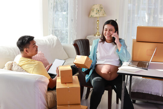 Asian couple working on box packaging, attractive young happy asian pregnant woman sitting and using mobile smart phone while writing order from customer with computer and husband working together