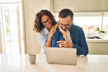 Middle age hispanic couple hugging each other using laptop at kitchen