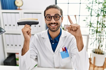 Young hispanic dentist man comparing teeth whitening smiling with an idea or question pointing finger with happy face, number one