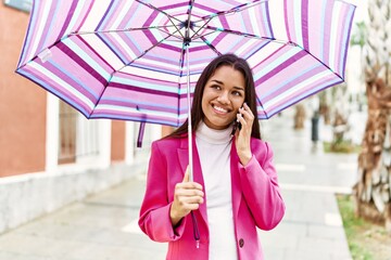 Young latin woman talking on the smartphone holding umbrella at street