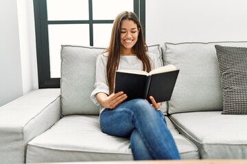 Naklejka premium Young latin woman reading book sitting on sofa at home