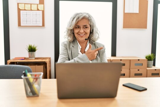 Middle age businesswoman sitting on desk working using laptop at office cheerful with a smile of face pointing with hand and finger up to the side with happy and natural expression on face