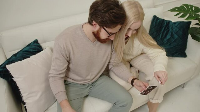 Millennial European couple looking at their baby ultrasound picture while sitting on a white sofa in their modern apartment. High quality 4k footage