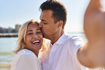 Fototapeta premium Middle age man and woman couple hugging each other and kissing make selfie by the smartphone at seaside