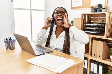 Black woman with braids working at the office speaking on the phone shouting and screaming loud to side with hand on mouth. communication concept.