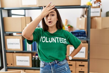 Young brunette woman wearing volunteer t shirt at donations stand surprised with hand on head for...