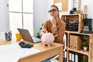 Young blonde woman at the office putting coin into piggy bank serious face thinking about question...