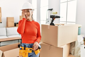 Middle age grey-haired woman wearing hardhat standing at new home covering one eye with hand, confident smile on face and surprise emotion.