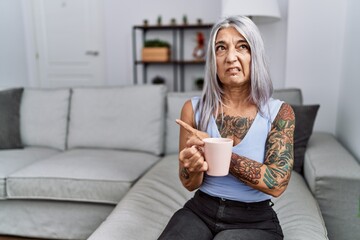 Middle age grey-haired woman drinking coffee sitting on the sofa at home pointing aside worried and...