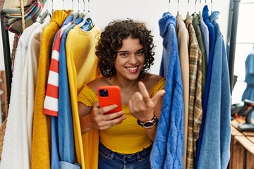 Young hispanic woman searching clothes on clothing rack using smartphone beckoning come here gesture with hand inviting welcoming happy and smiling