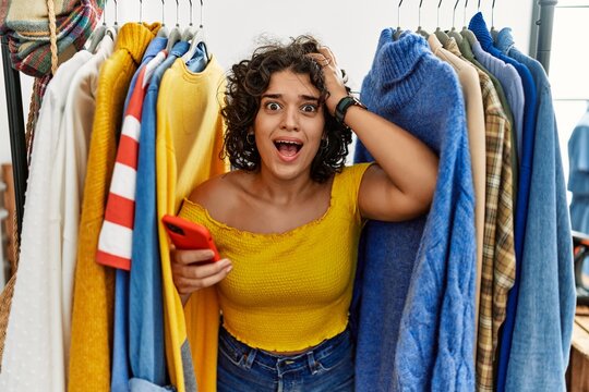 Young hispanic woman searching clothes on clothing rack using smartphone crazy and scared with hands on head, afraid and surprised of shock with open mouth