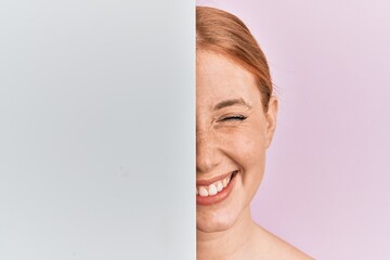 Young irish woman holding blank empty banner showing half face winking looking at the camera with...