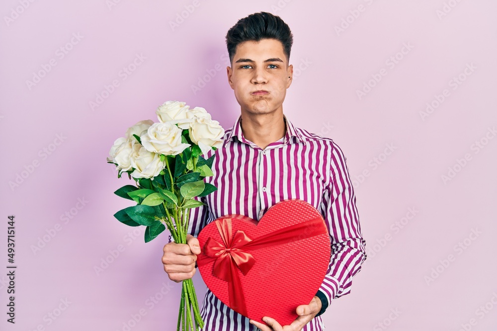 Canvas Prints Young hispanic man holding anniversary present and bouquet of flowers puffing cheeks with funny face. mouth inflated with air, catching air.