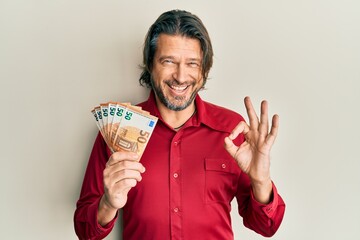 Middle age handsome man holding bunch of 50 euro banknotes doing ok sign with fingers, smiling friendly gesturing excellent symbol - obrazy, fototapety, plakaty