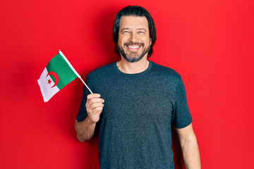 Middle age caucasian man wearing hijab holding algeria flag looking positive and happy standing and smiling with a confident smile showing teeth