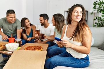 Group of young hispanic friends eating italian pizza sitting on the sofa. Woman smiling happy and using smartphone at home.