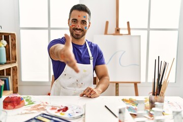 Young hispanic man with beard at art studio with painted face smiling friendly offering handshake as greeting and welcoming. successful business.