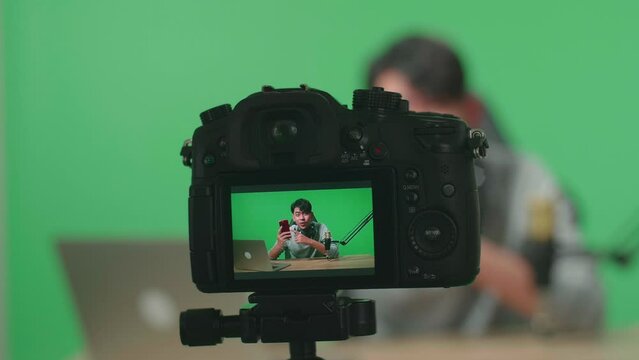 Close Up Of A Camera Monitor Recording Asian Man With Headphone And Computer Reviewing Mobile Phone On Green Screen Background
