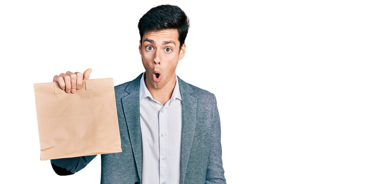 Young hispanic business man holding paper bag with bread scared and amazed with open mouth for surprise, disbelief face