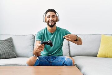 Handsome hispanic man wearing headphones playing video game holding controller smiling happy pointing with hand and finger