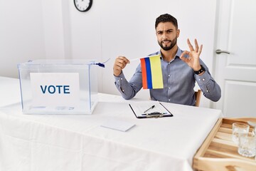 Young handsome man with beard at political campaign election holding colombia flag doing ok sign with fingers, smiling friendly gesturing excellent symbol