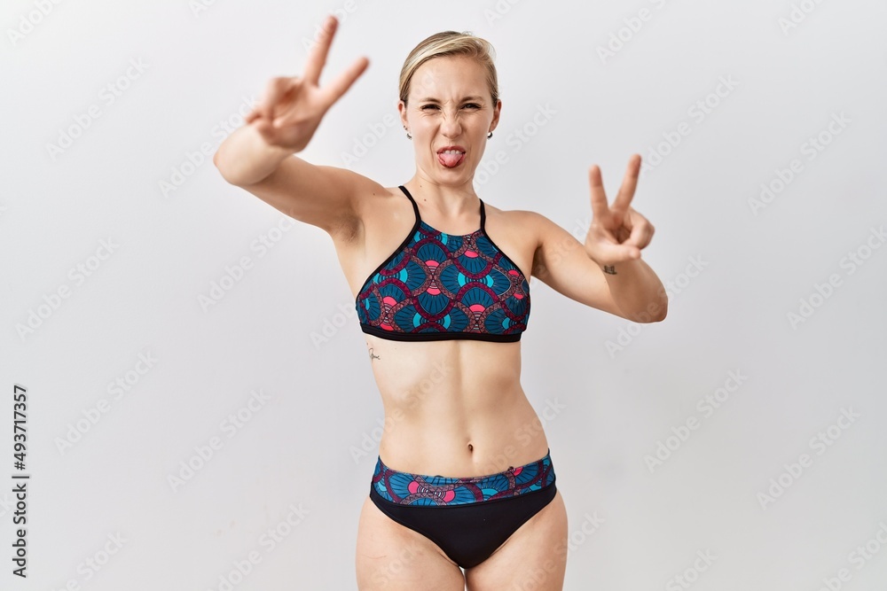 Poster Young blonde woman wearing sporty bikini over isolated background smiling with tongue out showing fingers of both hands doing victory sign. number two.