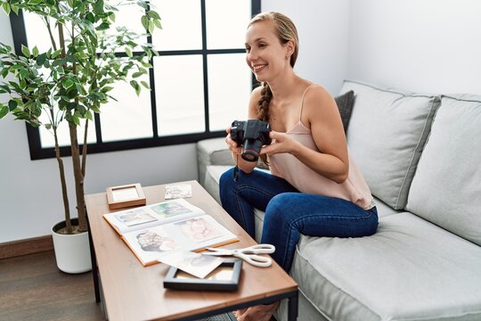 Young caucasian woman making photo book holding camera sitting on sofa at home