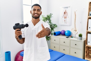 Naklejka na ściany i meble Young indian physiotherapist holding therapy massage gun at wellness center smiling friendly offering handshake as greeting and welcoming. successful business.