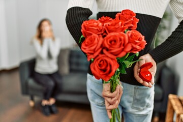 Man suprising woman with bouquet of roses and engagement ring at home.