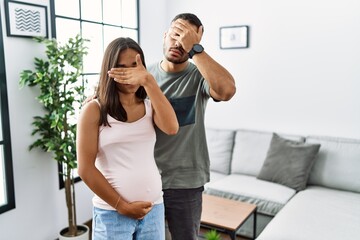 Young interracial couple expecting a baby, touching pregnant belly covering eyes with hand, looking serious and sad. sightless, hiding and rejection concept