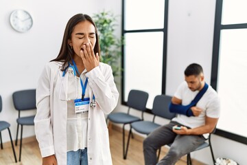 Young asian doctor woman at waiting room with a man with a broken arm bored yawning tired covering mouth with hand. restless and sleepiness.
