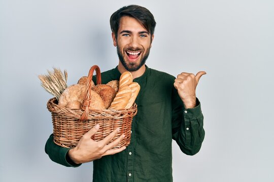 Young hispanic man holding wicker basket with bread pointing thumb up to the side smiling happy with open mouth