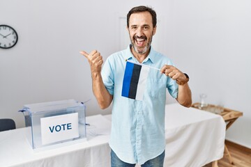 Middle age man with beard at political campaign election holding estonia flag pointing thumb up to the side smiling happy with open mouth