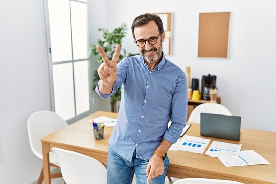 Middle age hispanic man with beard wearing business clothes at the office smiling looking to the camera showing fingers doing victory sign. number two.