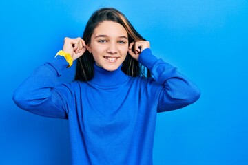 Young brunette girl wearing turtleneck sweater smiling pulling ears with fingers, funny gesture....
