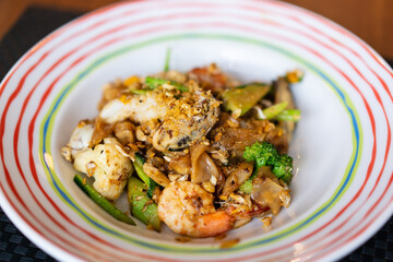 stir fried wide noodles with seafood or fried rice noodles with seafood soy sauce in white colorful rim plate on top view wooden table and placemat which consists of mussel shrimp squid and vegetable