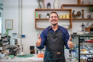 Portrait of smiling owner man standing at his cafe with thumbs up hand. Coffee owner standing with apron in coffee shop to welcome customer.