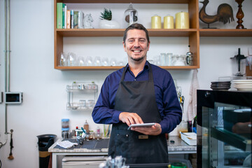 Portrait of smiling owner man standing at his cafe. Coffee owner standing with apron in coffee shop to welcome customer. - 493723956