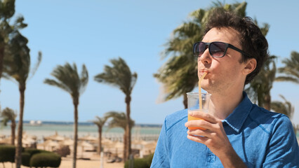 Man on vacation backdrop of palm trees, sea, drinks orange cocktail, enjoying summer holiday at all-inclusive beach resort. Guy in sunglasses, blue tennis shirt on azure sea with sun umbrellas sunbeds