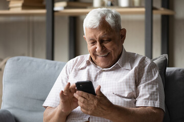 Happy elderly man look at cell phone screen rest on sofa at home, smiling enjoy received message...