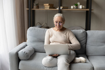 Senior woman in glasses rest on sofa with laptop, make order, buy goods, electronic services on internet via retail websites. Older generation use modern tech, websurfing, spend leisure online concept